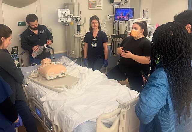 Medical team gathered around a hospital bed in a clinical room, practicing procedures on a training mannequin. The scene conveys focus and teamwork.