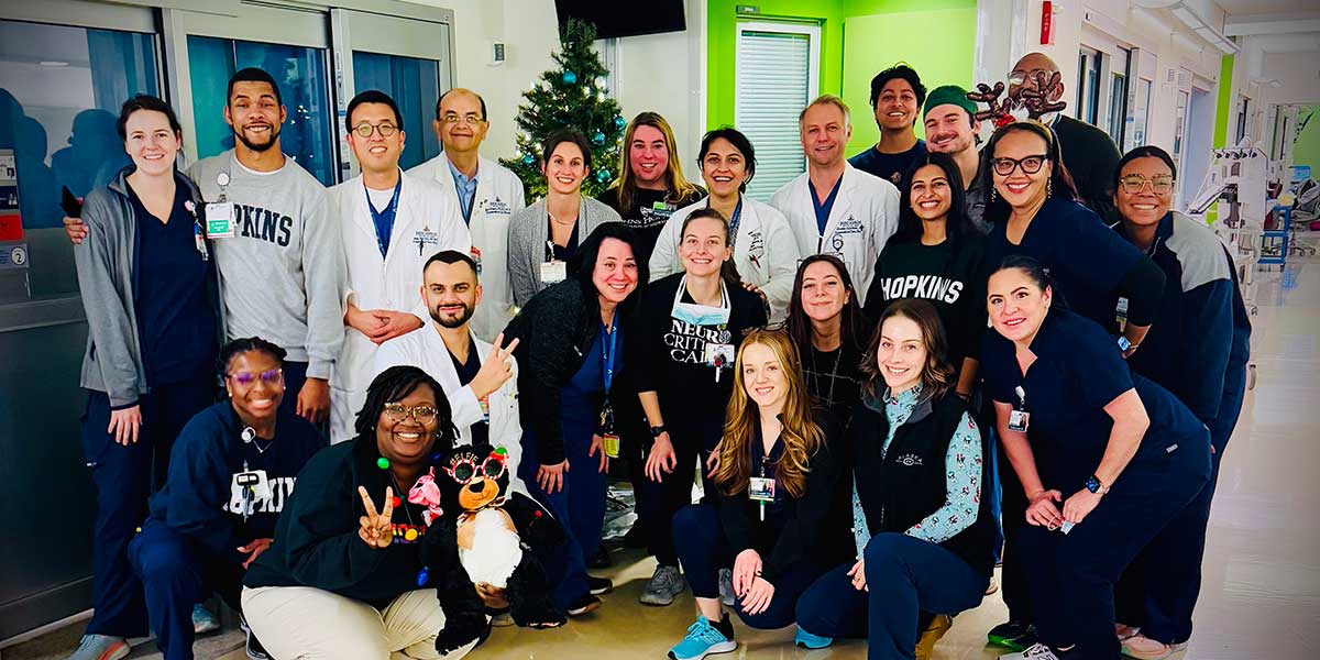 A diverse group of 21 healthcare professionals pose smiling in a hospital hallway, with festive holiday decorations like a small Christmas tree visible.