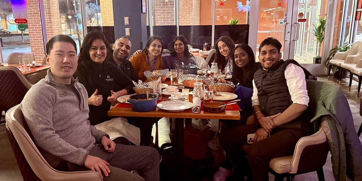 A group of eight neurocritical care fellows sit around a table at a cozy restaurant, smiling and enjoying a meal together. The warm lighting and contemporary decor create an inviting atmosphere.