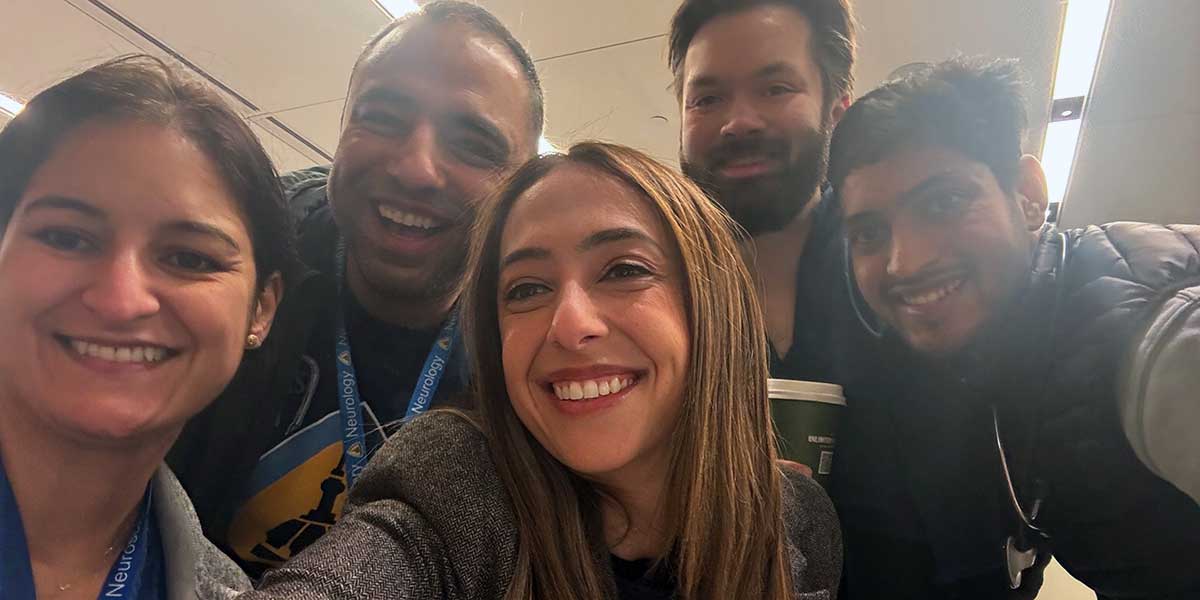 A group of five neurocritical fellows smiling in a well-lit indoor setting. They appear happy and engaged, suggesting a friendly, casual gathering.