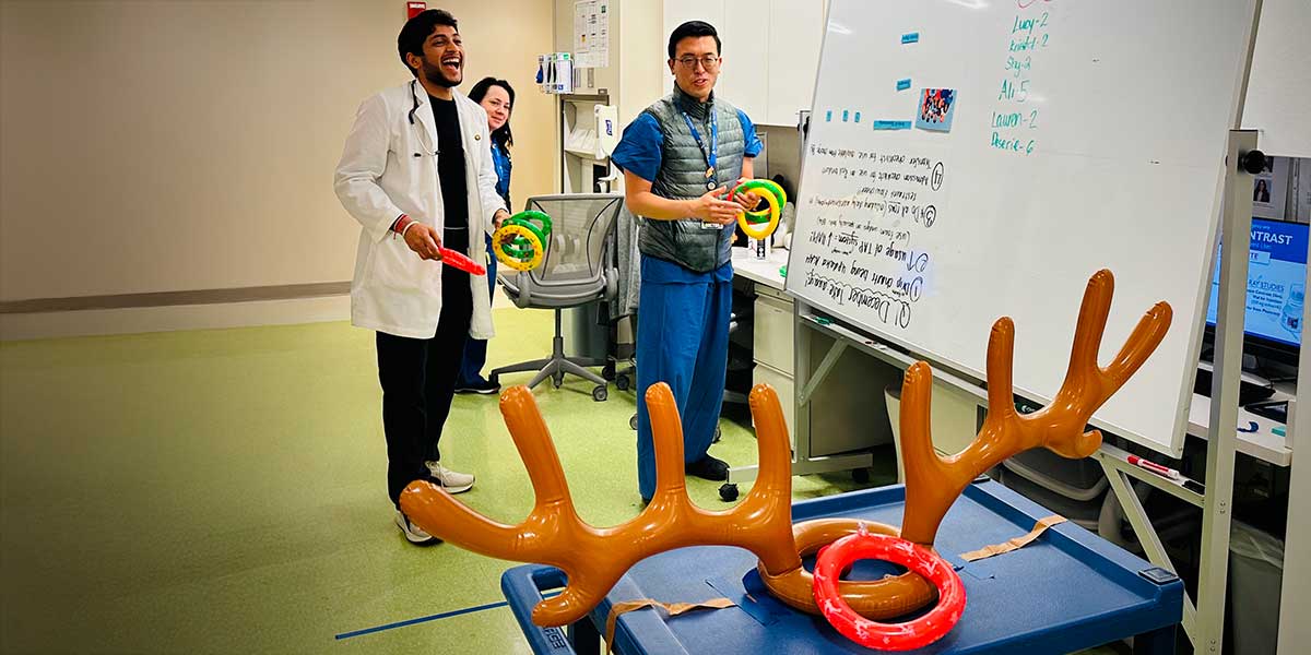 Neurocritical fellows play a fun ring toss game on antler-shaped targets in a medical office space, smiling and relaxed. A whiteboard with game scores in the background.