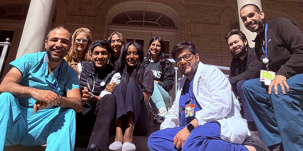 A group of smiling neurocritical fellows and faculty in scrubs sit on steps outside a brick building with columns, enjoying a sunny day, exuding camaraderie.