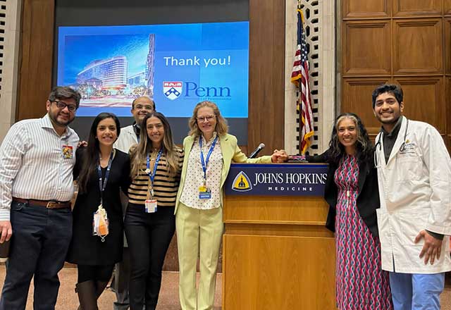 A group of NCCU faculty and trainees stand together smiling in front of a lectern with a Johns Hopkins Medicine sign, a presentation screen behind them. The setting appears professional and the mood is positive and collaborative.