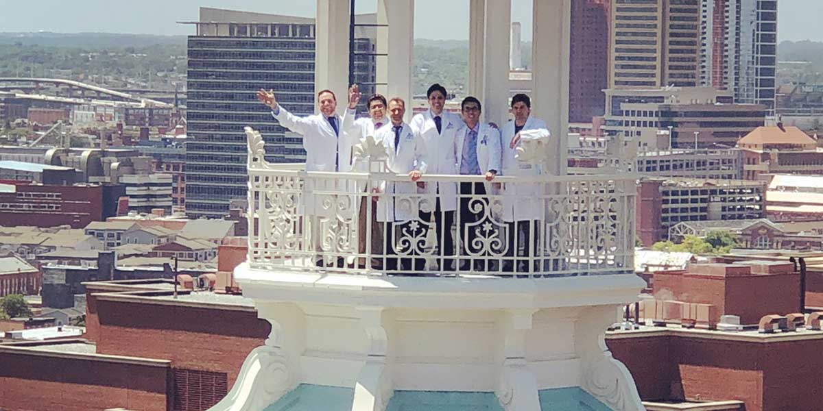 NCCU fellows in white lab coats waving as they stand on the dome balcony of the Johns Hopkins Billings building