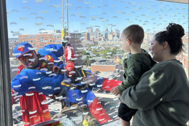 A young child being held up by an adult watching a window washer dressed in superhero costume 