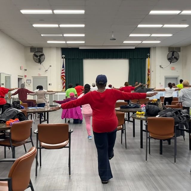 Photo of a woman in a class room walking down the room with her arms open, other class mates in the room.