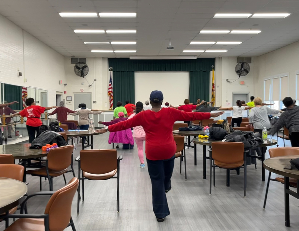 Photo of a woman in a class room walking down the room with her arms open, other class mates in the room.