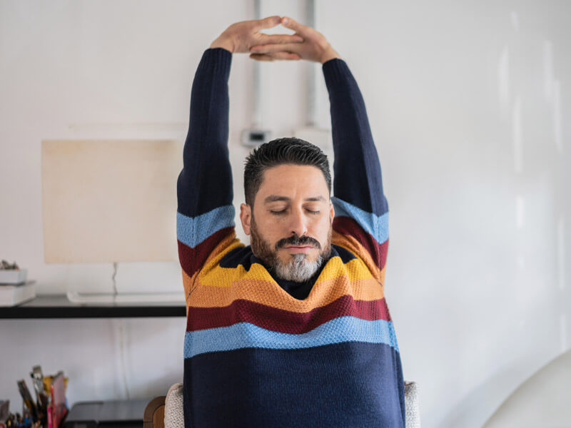 Man stretches his back and arms while sitting at his work desk