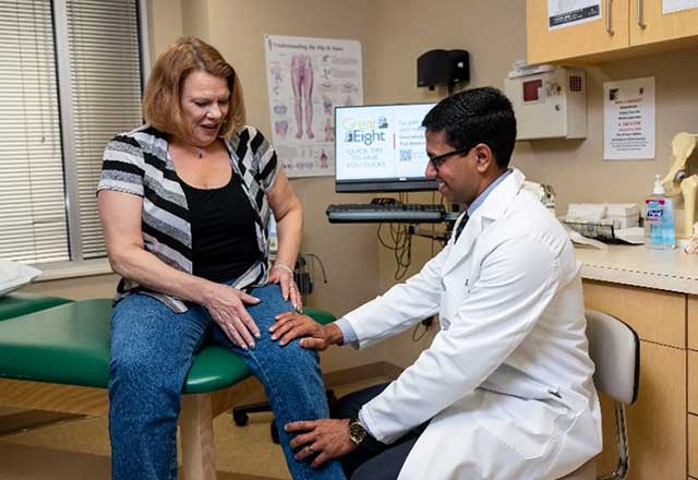 A doctor in a white coat examines a woman's knee in a medical office. The woman looks concerned. Posters and a computer are visible in the background.