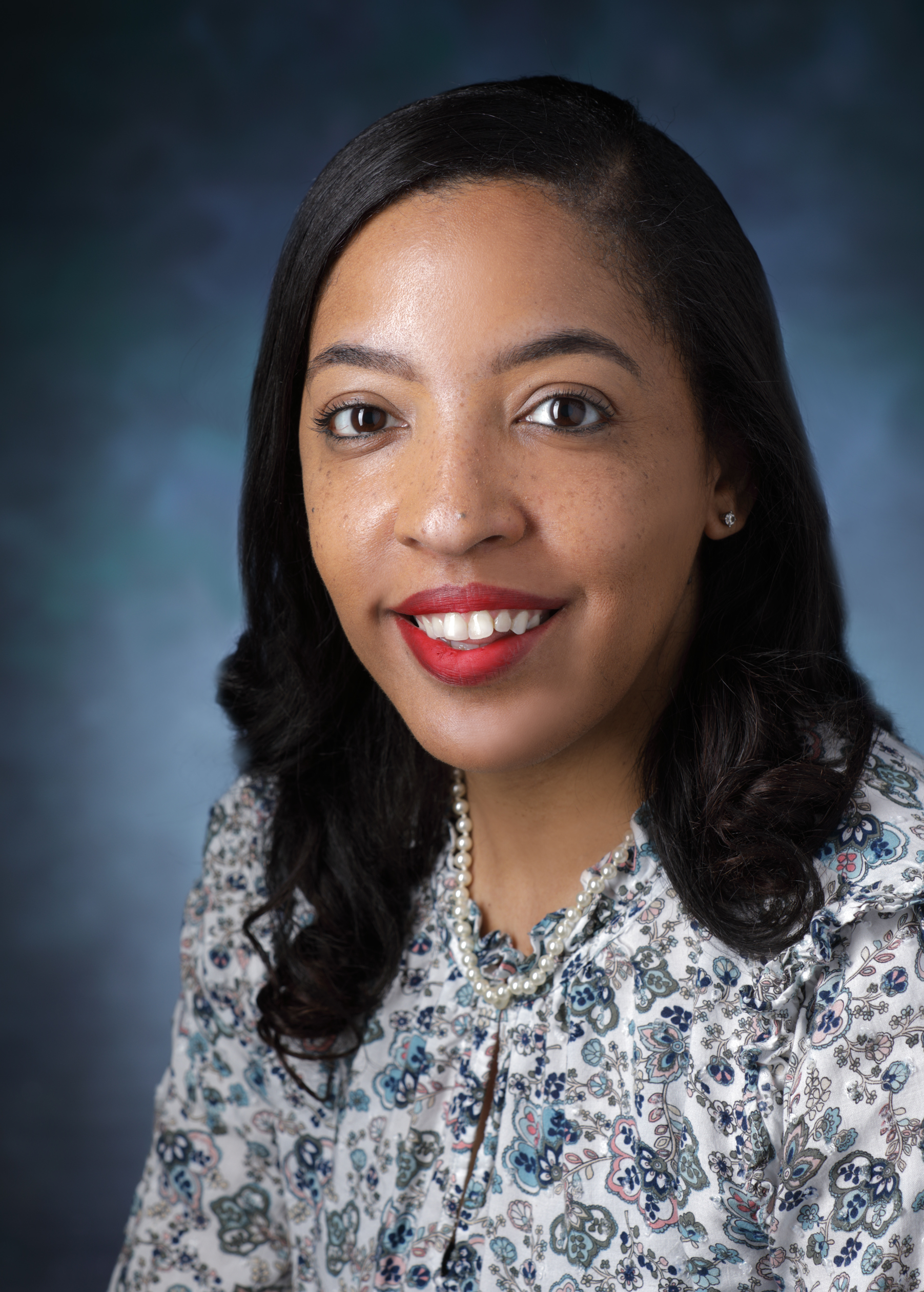 Andrea Mansfield, wearing a blue and white flower print blouse, smiles in front of a blue photo backdrop.