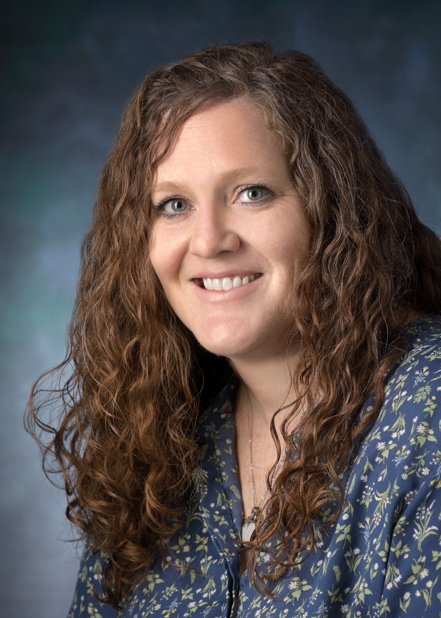 Lesa Miller, wearing a blue flower print blouse, smiles in front of a blue photo backdrop.