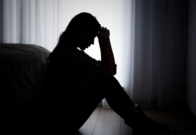 A silhouette of a woman sitting at the foot of a bed, with her hands on her face.