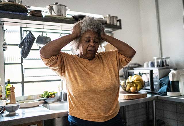 A older women in her kitchen holding her head in distress.