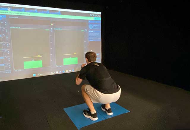 Patient using muscle performance assessment technology, standing on a plate and squatting in front of a screen reading out measurements.