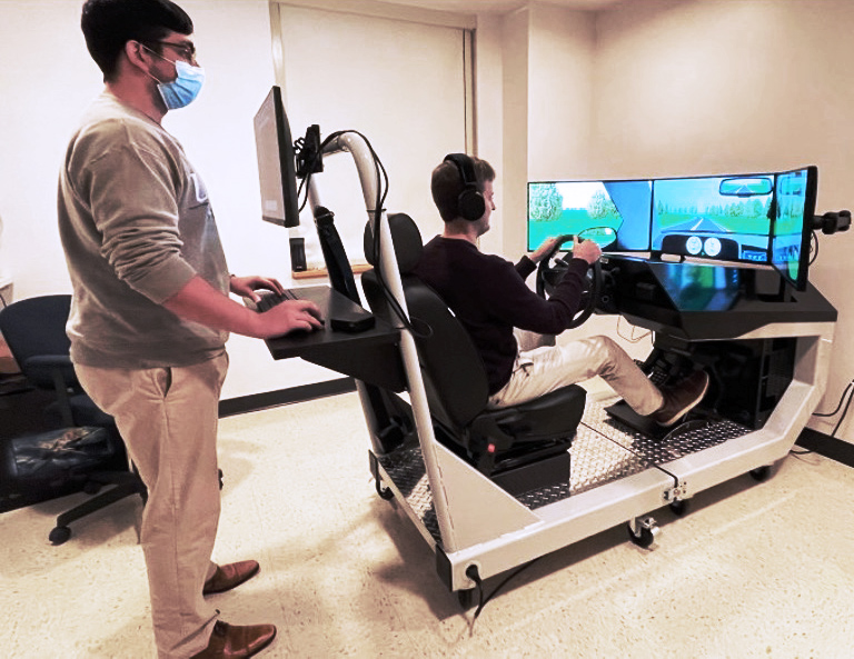 A person sitting in a driving simulator and a person at the back of a driving simulator. Photo courtesy of Tory Spindle.