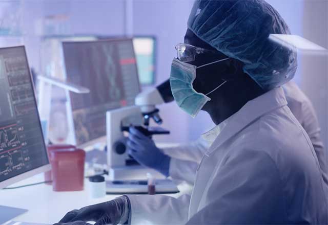 Healthcare researcher wearing a mask and hair net, looking into a microscope.