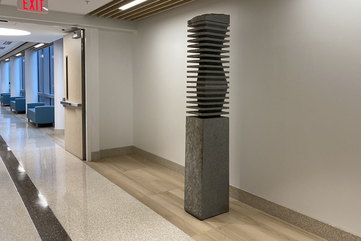 Photograph of a sculpture created by Jesús Moroles. The sculpture is gray stone pillar with an undulating design at the top, cross-sectioned by distinct lines jutting out at the same dimensions as the lower base.
