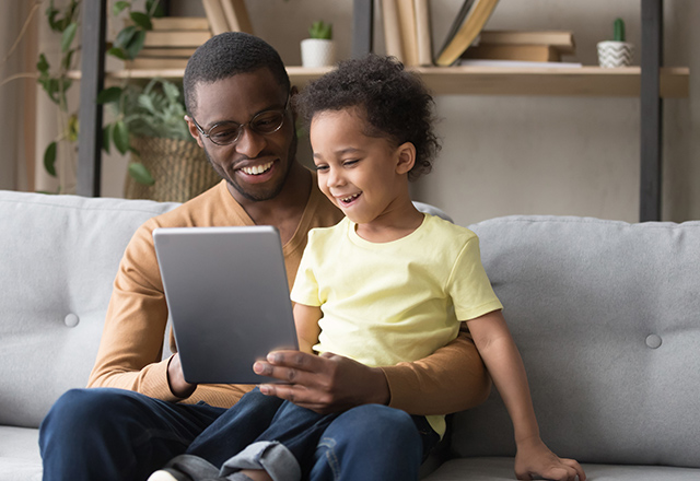 Excited loving african American dad hold tablet sit on couch with cute preschooler son watch cartoon together, smiling happy black dad and small boy kid relax on sofa at home play game on pad