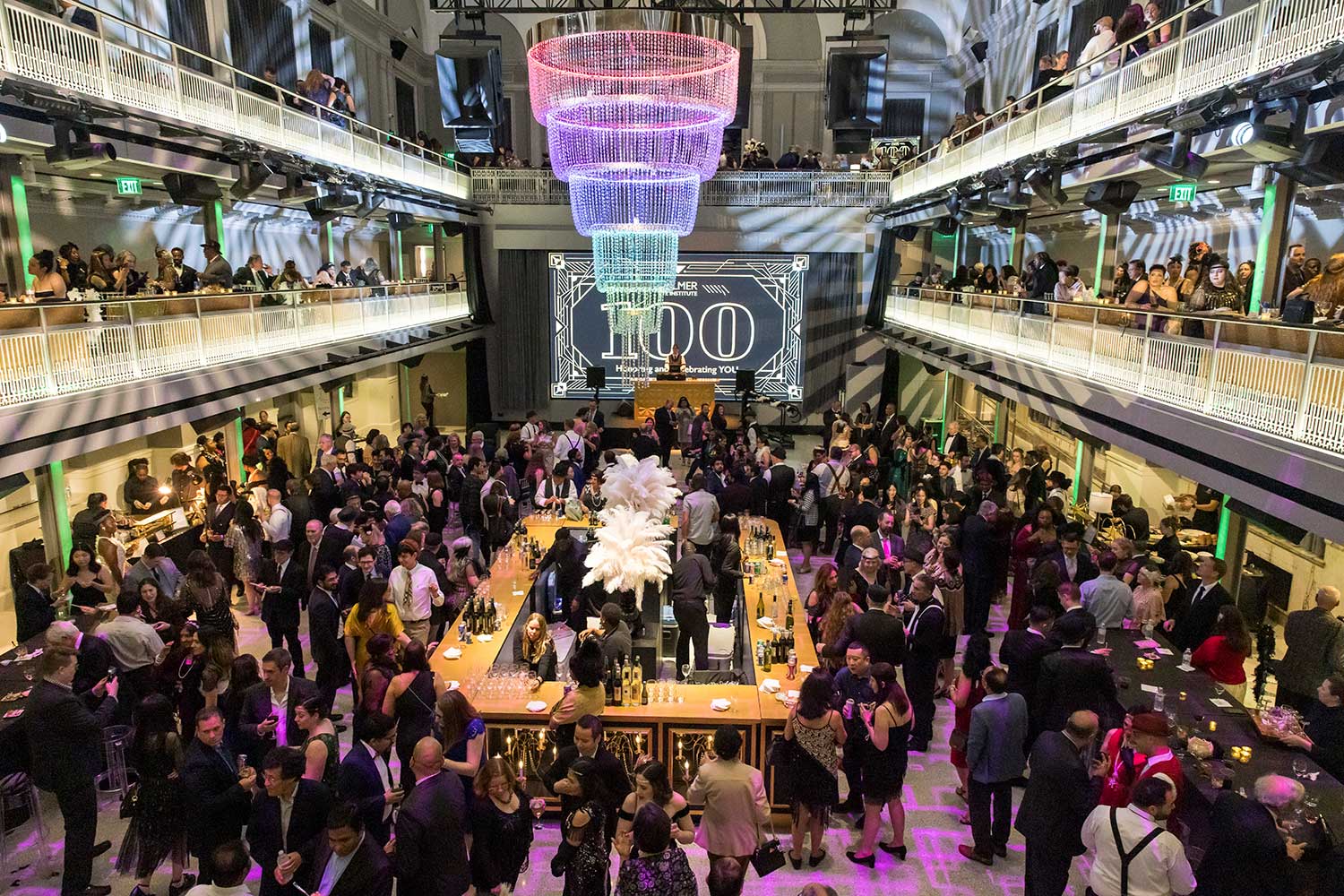 Crowded ballroom with large chandelier. In the background, a bright sign reads 100