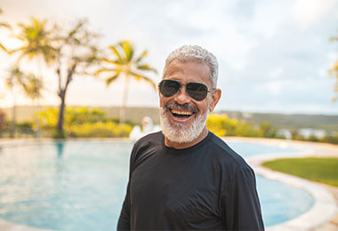 An older man laughs in front of a pool with a palm tree off to the side. He is wearing sunglasses to protect his eye health.
