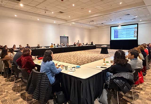 A large panel sits around a U shaped table with a large presentation screen in the middle