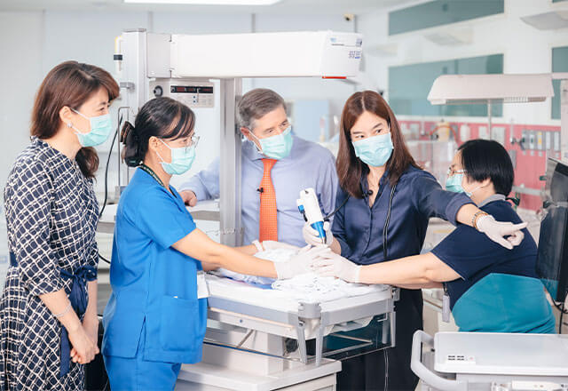Neil Bressler, center, discusses retina images being obtained in a premature infant by Atchareeya Wiwatwongwana, right center, while the baby is soothed by Benja Yatikul, far right, (the head nurse in the ROP clinic), and Kwanhathai Kantaroj, left center (a neonatal nurse specialist), as Jun Kong, far left, prepares communication with retina experts in Shanghai