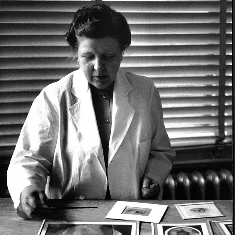 Black and white photo of Annette Smith Burgess wearing a white coat, examining printed illustrations on a desk