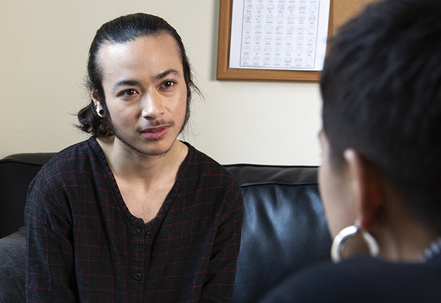A genderqueer person sits on a couch, talking to a healthcare provider.