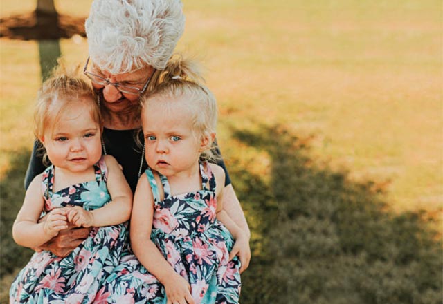 A senior looks down at the two toddler-age girls sitting in her lap. The girls wear matching floral dresses. The sun casts a warm glow on the grassy lawn behind them.