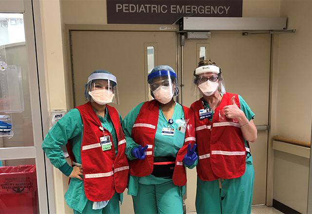 Three Pediatric staff members pose in front of the Pediatric Emergency Room doors. They are wearing protective equipment; one of them flashes a pair of peace signs while another gives a thumbs-up.
