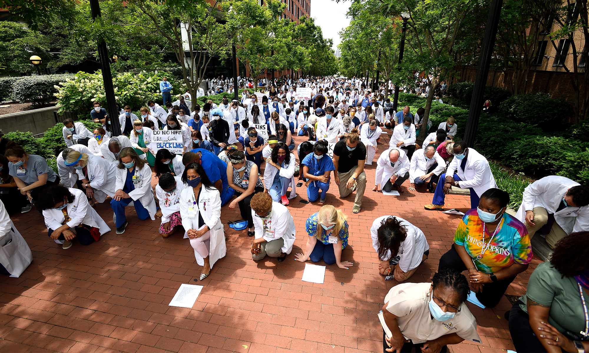 Hundreds of Johns Hopkins clinicians in white lab coats kneel on the campus of the Johns Hopkins hospital.