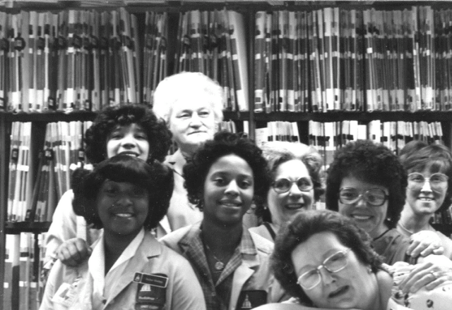 A vintage picture of a group of radiology film clerks smiling in front of the stack of films.