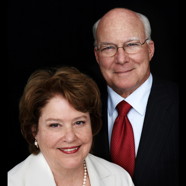Michael and Anne Armstrong, in a formal portrait with a black background. Michael is wearing a dark suit, white button down ad red tie, Anne is wearing an off-white coat, pearl necklace and pearl earrings with a white top.