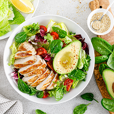 Grilled chicken salad with tomato, avocado, lettuce and spinach in a white bowl on a gray table