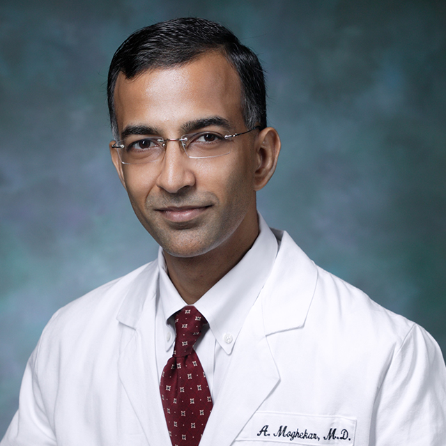 Neurologist Abhay Moghekar, in a formal portrait, wearing a white lab coat, white button down shirt and red tie.