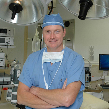 Martin Paul, wearing scrubs and a surgical cap, poses in an operating room, with various equipment and screens behind him