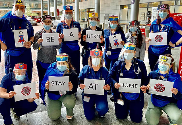 Johns Hopkins Children's Center staff in protective equipment pose together holding encouraging signs that read 'Be a hero, stay at home.' Each of the staff wear a different superhero's logo on their masks.