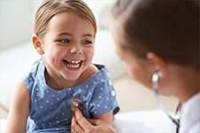 Una doctora ausculta el corazón de una niña que sonríe divertida.