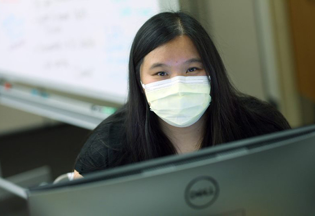 Tina Hoang sitting behind a computer screen, wearing a mask.