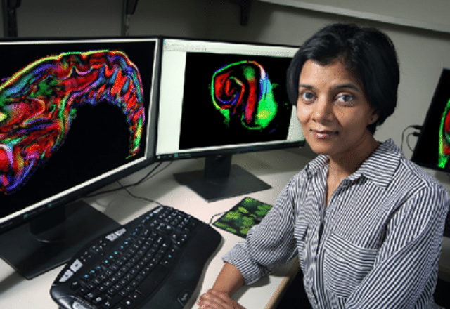 Manisha Aggarwal sitting at a desk with brain scans on two computer screens behind her. 
