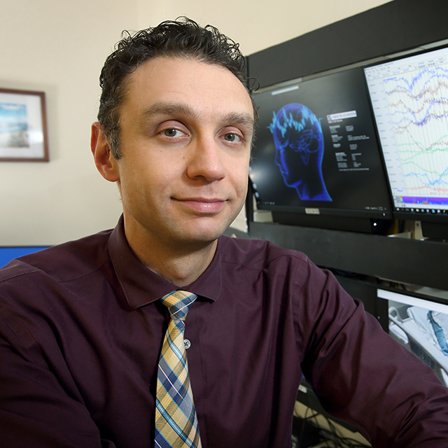 Ahmad Marashly, wearing a purple shirt and plaid tie, poses in front of computer monitors showing brain activity 